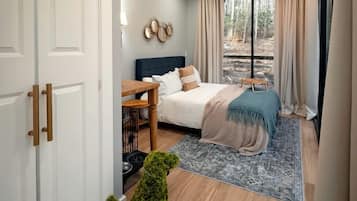 Ein Schlafzimmer mit einem Bett, einem kleinen Tisch, einer Zimmerpflanze und Blick ins Freie.