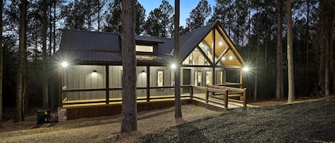 Una cabaña con terraza de madera, ventanas amplias y techo metálico, rodeada de árboles.