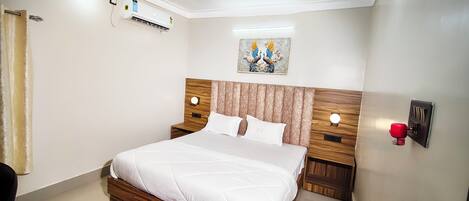 A hotel room with a bed, an air conditioning unit, a wall-mounted light, and a painting.