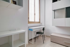 Ein Zimmer mit weißem Schreibtisch, einem Stuhl, einem Heizkörper, einem Fenster und einem eingebauten Schrank.