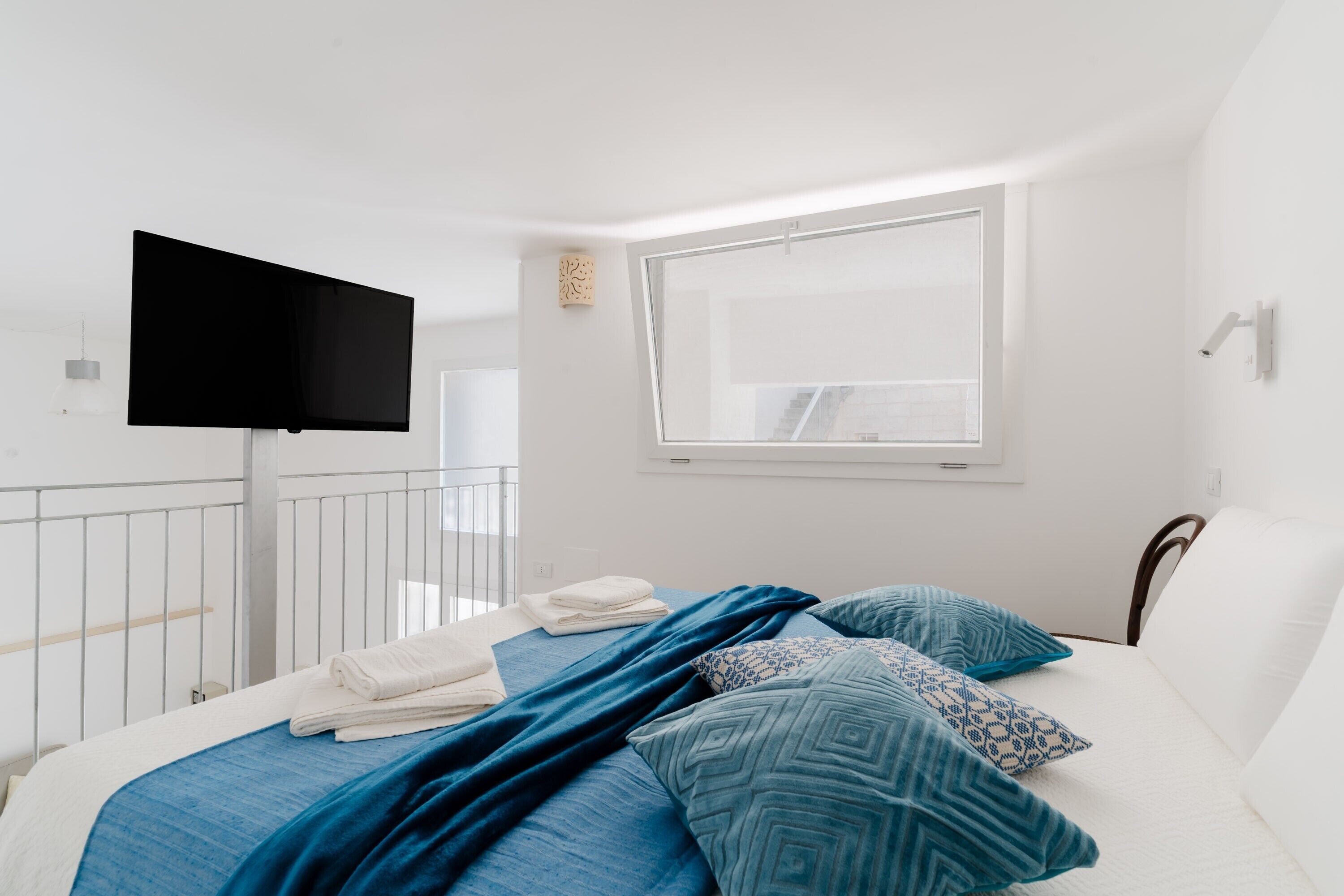 A bedroom with a bed, a flat-screen TV, a window, and a balcony railing.