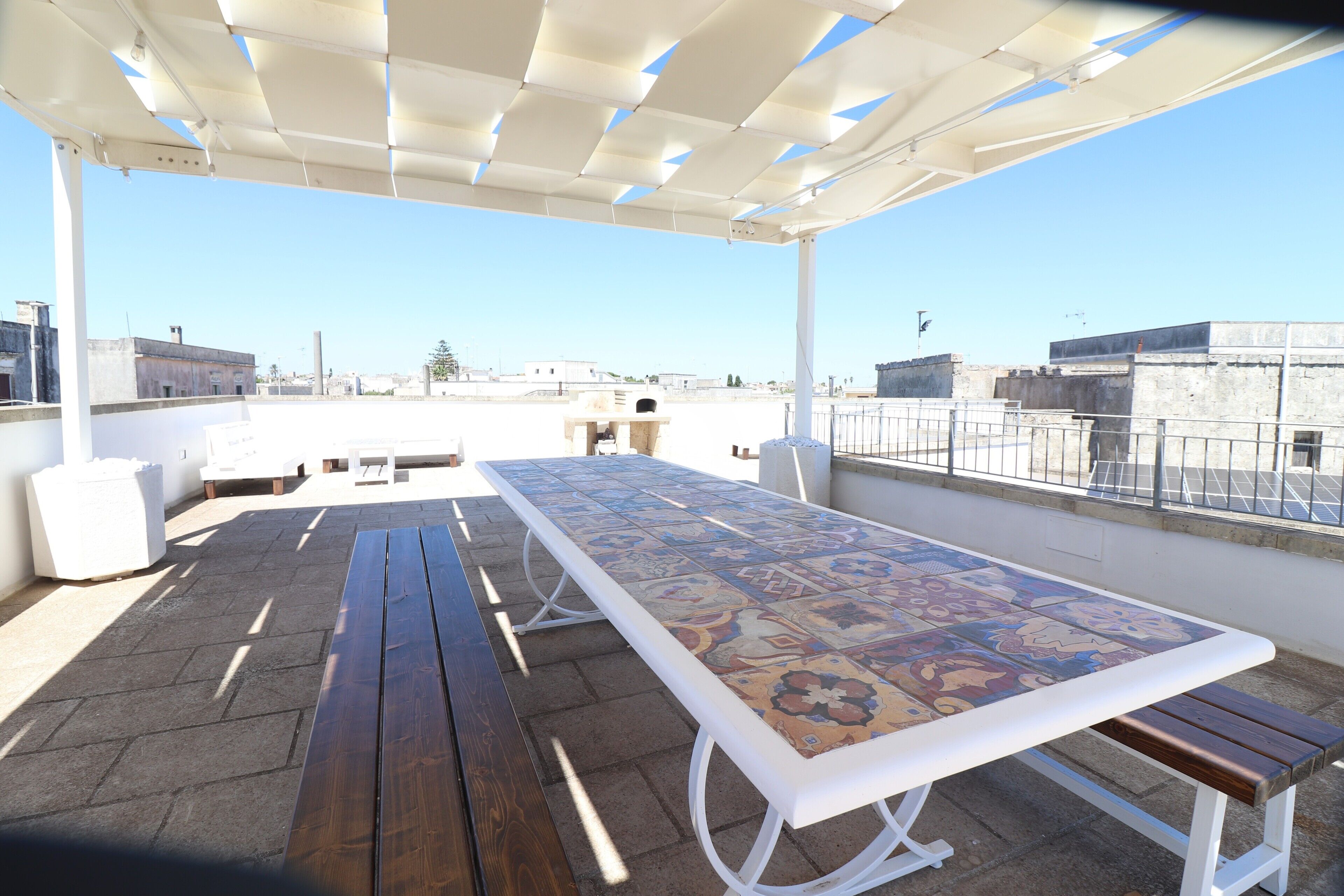 An outdoor patio with a long table and benches, a pergola overhead, and a view of the cityscape.