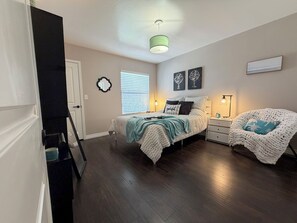 Ein Schlafzimmer mit Bett, Nachttischen, einem Sessel, einem Fenster mit Jalousien, einer Wanduhr und einer Deckenleuchte.