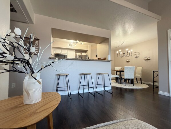 A modern kitchen with a central island, bar stools, a dining area with a round table, and a decorative vase with branches. - Serene & Secure Condo | Gated Community, Pool, Gym, and Dedicated Parking (Henderson)