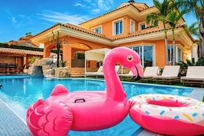 Una piscina con flotadores de flamenco rosa y anillos de rosquilla, un edificio de hotel con techo de tejas y sillas de descanso.