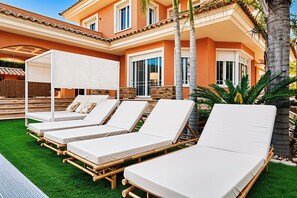 Zona de piscina con sillas de descanso, un área de estar cubierta y un edificio con grandes ventanales y techo de tejas.