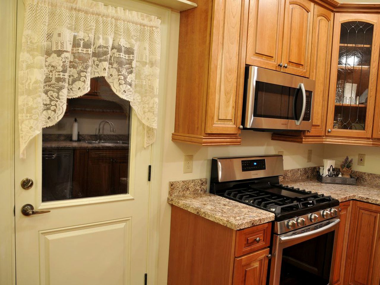A kitchen with wooden cabinets, a stainless steel microwave, a gas stove, and a window with a lace curtain.