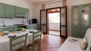 Une cuisine avec des meubles de couleur verte, une table à manger avec des chaises, un téléviseur et une porte-fenêtre coulissante donnant sur un espace extérieur.