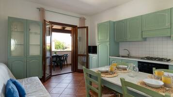 A room with a bed, a dining table set for two, and a kitchen area with green cabinets and a tiled backsplash.