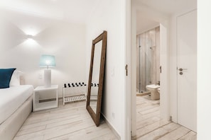 Habitación de hotel con cama, mesita de noche, lámpara, espejo y vistas al baño.