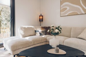 Un salon moderne avec un canapé d’angle, une table basse, un lampadaire et une grande fenêtre donnant sur la forêt.