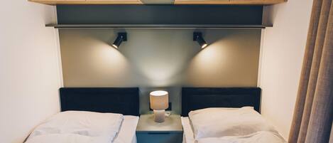A bedroom with two beds, a nightstand with a lamp, wooden cabinets above, and a curtain on the right.