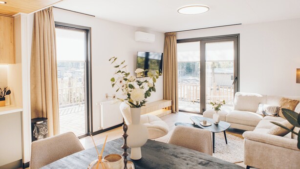 A modern living room with a large window, a flat-screen TV, and a dining area with a vase of flowers.