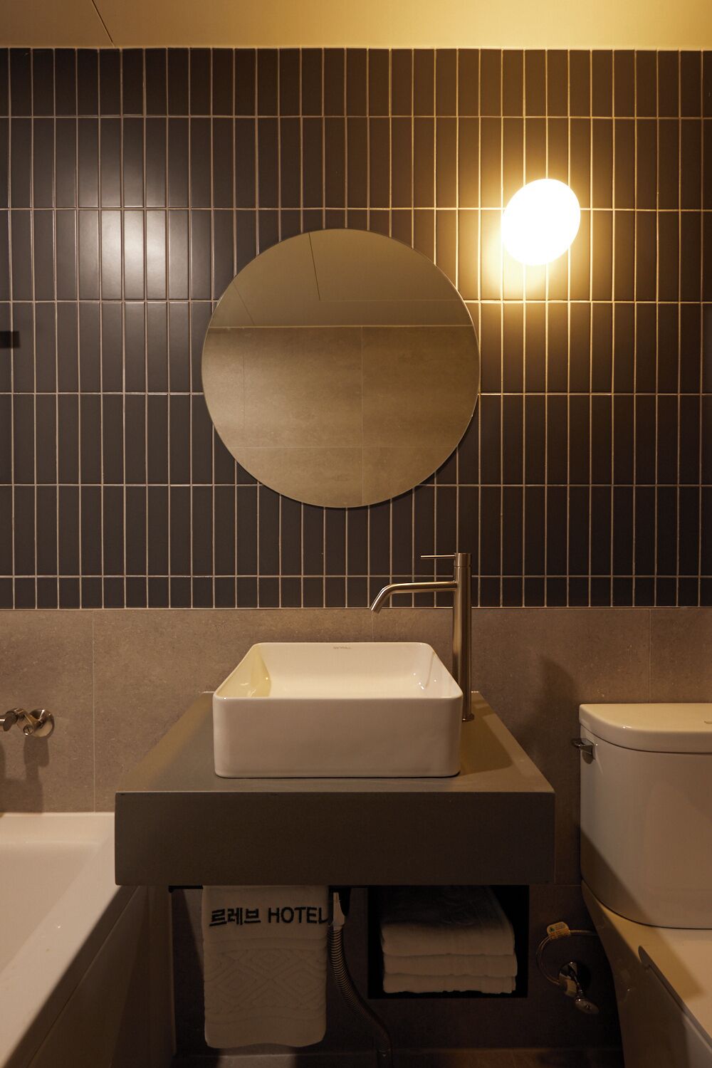 A bathroom with a round mirror, a rectangular sink, a toilet, and a towel dispenser.