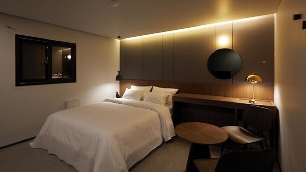 Una habitación de hotel moderna con cama, mesita de noche, lámpara y un pequeño rincón de descanso.