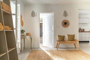 Ein Zimmer mit einer hölzernen Bank, einer weißen Tür, die nach draußen führt, einem gewebten Teppich und einem an der Wand befestigten Dekorationselement.