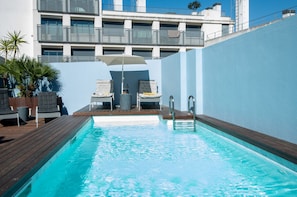 A swimming pool with lounge chairs and an umbrella, surrounded by a blue wall and a building with balconies. - Lbb1 (Lisbon)