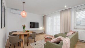 Ein modernes Wohnzimmer mit einem runden Holztisch, zwei grauen Sesseln, einem grünen Sofa, einer pinkfarbenen Tagesdecke und einer hängenden Pendelleuchte.