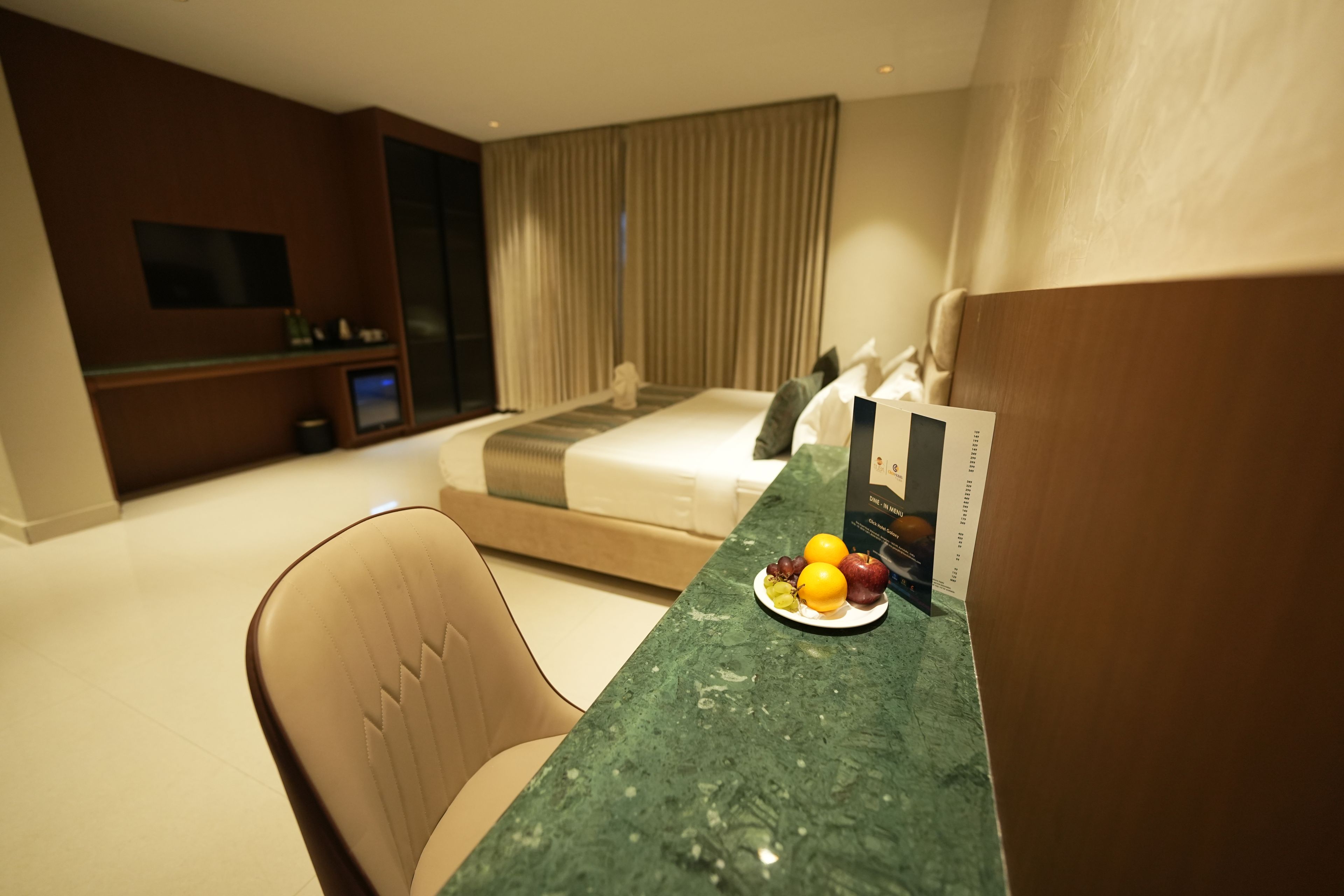A hotel room with a bed, a desk with a plate of fruit, a chair, a television, and a minibar.
