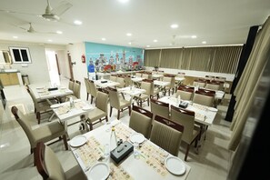 A spacious dining area with multiple tables and chairs, a cityscape mural on the wall, and ceiling fans. - Galaxy Air Suites (Bengaluru)
