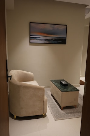 Una habitación con un sillón color beige, una mesita con un control remoto y un cuadro en la pared.
