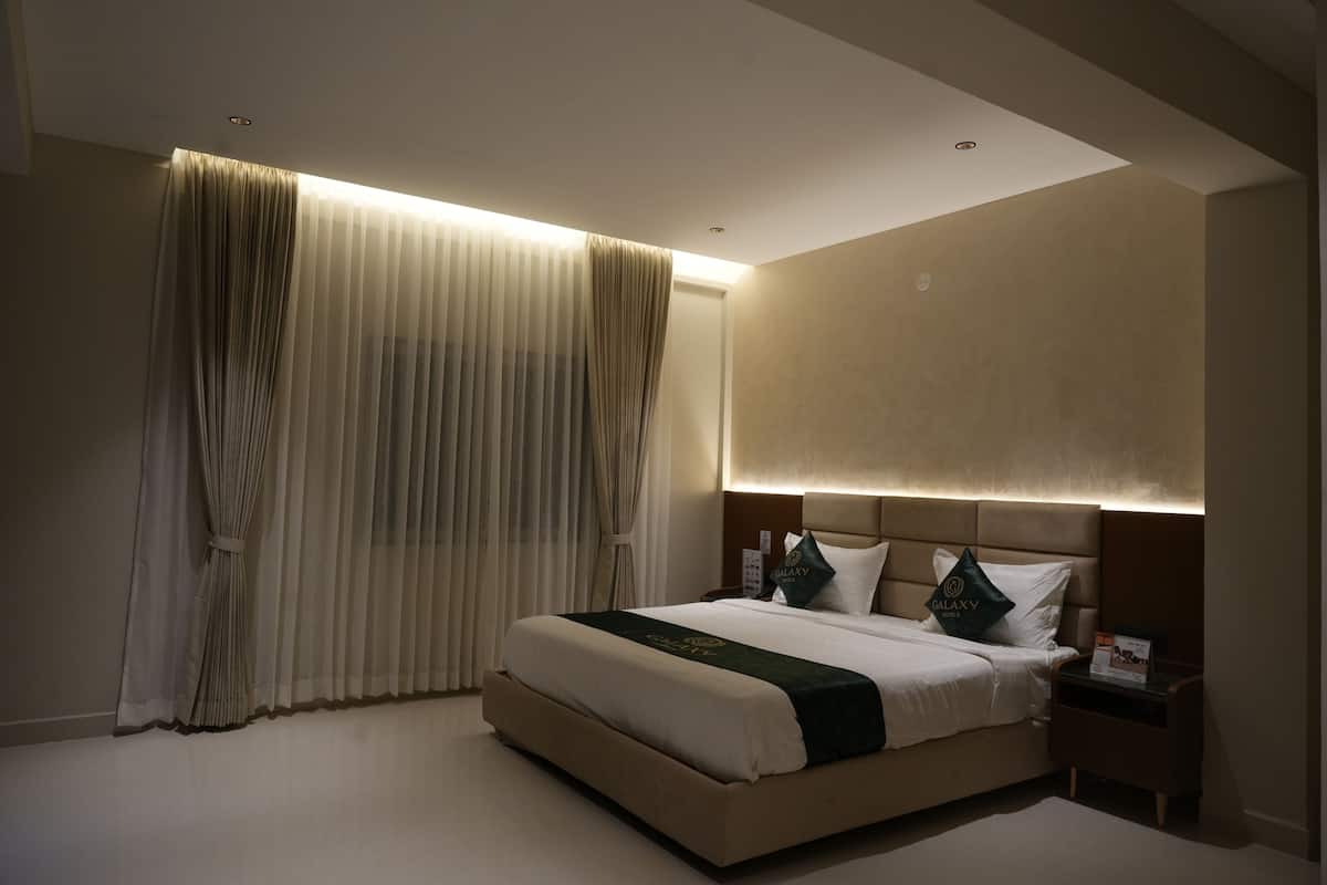 A modern hotel room with a large bed, a headboard with integrated lighting, a bedside table, and a large window with curtains.
