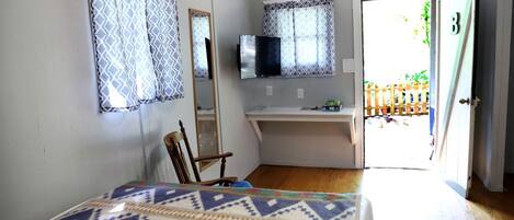A bedroom with a bed, a chair, a desk, a TV, and a door leading outside.