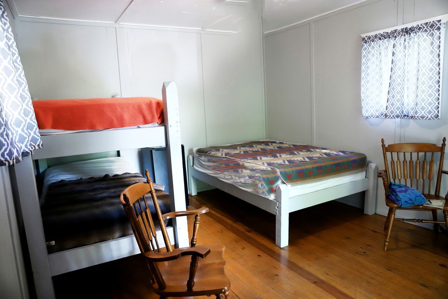 A dorm-style room with bunk beds, a wooden chair, and a window with patterned curtains.