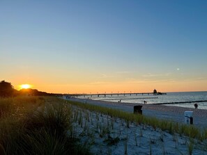 Ein Strand mit einem Steg, der sich ins Wasser erstreckt, grasbewachsenen Dünen und einem Sonnenuntergang.