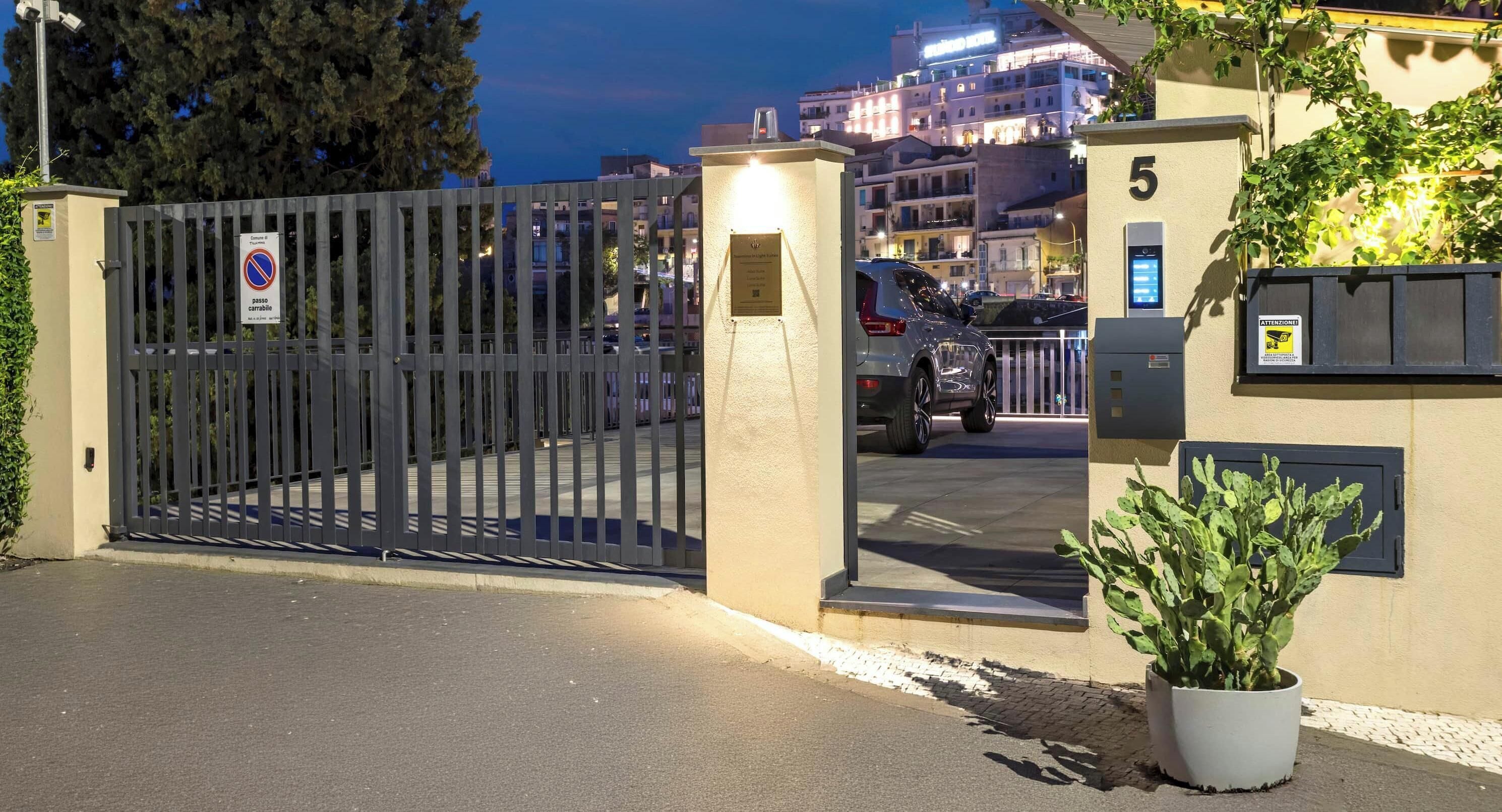 Un cancello chiuso dà accesso a un edificio con una pianta in vaso, un segnale che indica divieto di parcheggio e un edificio sullo sfondo con finestre illuminate.