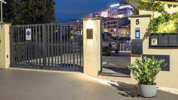 Un cancello chiuso dà accesso a un edificio con una pianta in vaso, un segnale che indica divieto di parcheggio e un edificio sullo sfondo con finestre illuminate.