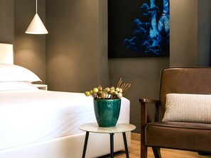 A hotel room with a bed, a small round table with a vase of dried flowers, a brown leather chair, and a painting on the wall. - Alba Suite-Taormina In Light Suites (Taormina)