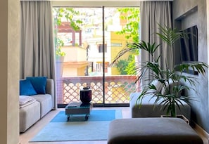 A modern living room with a sofa, a coffee table, a TV, and a large window with a view of buildings and greenery. - Alba Suite-Taormina In Light Suite (Taormina)