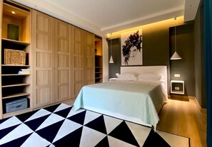 A bedroom with a bed, a geometric patterned rug, a wardrobe, shelves with items, and a wall art piece. - Alba Suite-Taormina In Light Suite (Taormina)