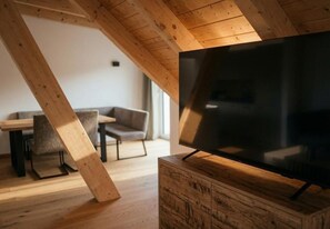 Uma sala de estar moderna com uma televisão de tela plana grande sobre um suporte de madeira, uma área de jantar com mesa e cadeiras, e teto inclinado com vigas de madeira aparentes.