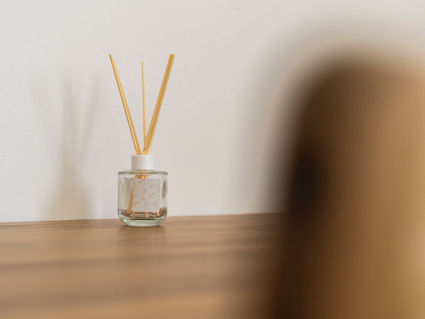 A clear glass bottle with reed sticks on a wooden surface against a plain wall.