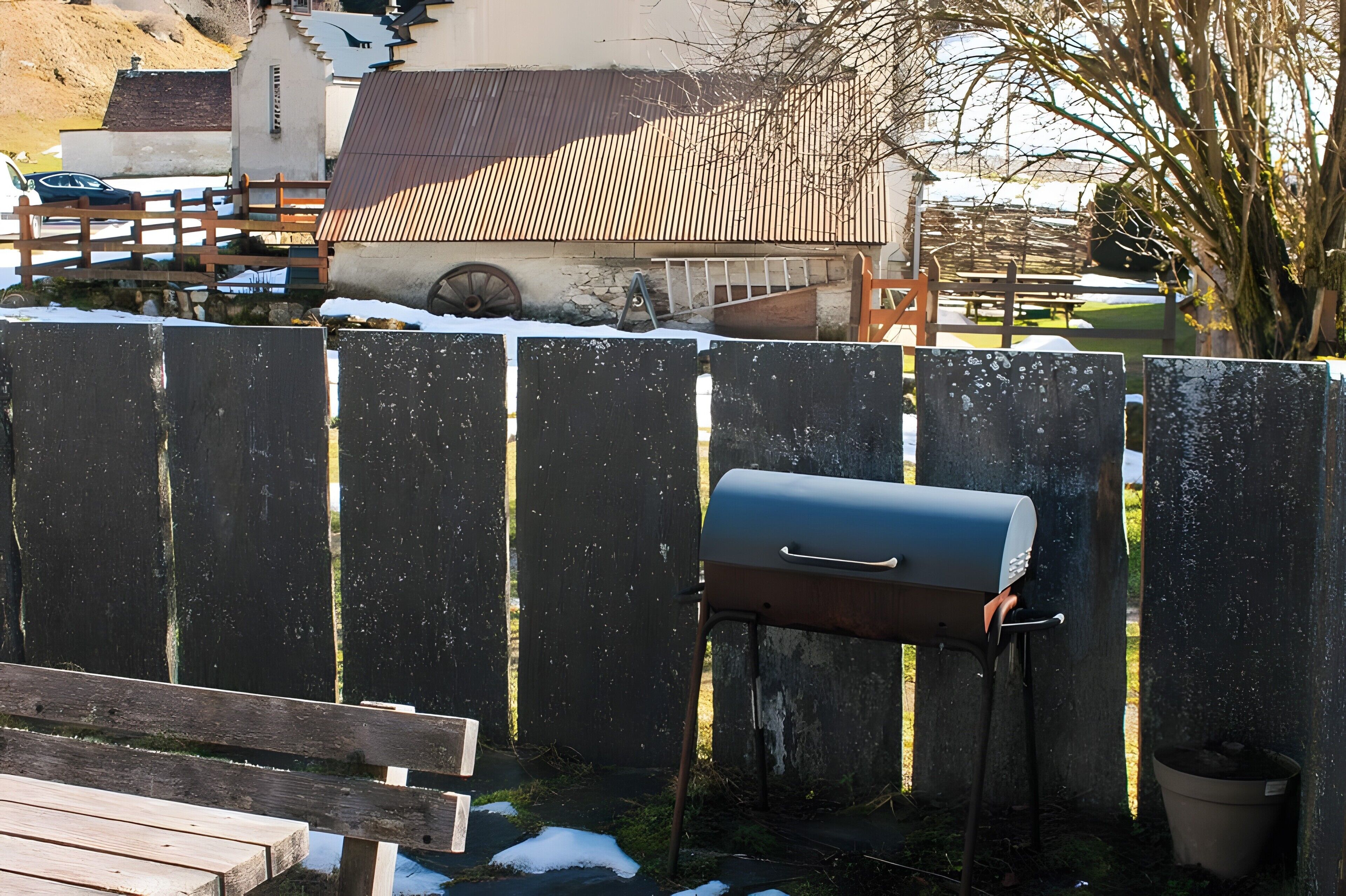 A mailbox attached to a wooden fence, a bench, and a building with a sloped roof in the background.