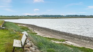Vista del río con un banco de madera, orilla pedregosa y edificios a lo lejos.