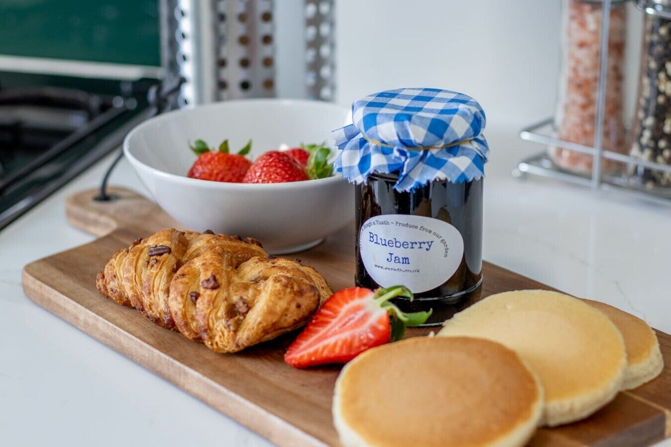A wooden board with pastries, a jar of blueberry jam, and a bowl of strawberries.