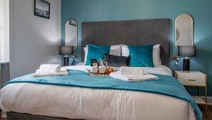 Een net opgemaakt bed met een blauwgroene deken, een nachtkastje met een lamp en een dienblad met toiletartikelen.