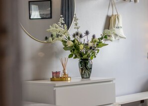 Ein weißer Nachttisch mit einer Glasvase, die weiße Blumen, eine kleine Kerze und ein dekoratives Objekt enthält.