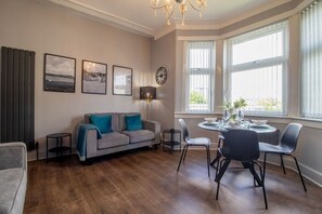 Ein Wohnzimmer mit grauem Sofa, einem Esstisch mit Stühlen, einer Stehlampe und einem großen Fenster mit Jalousien.