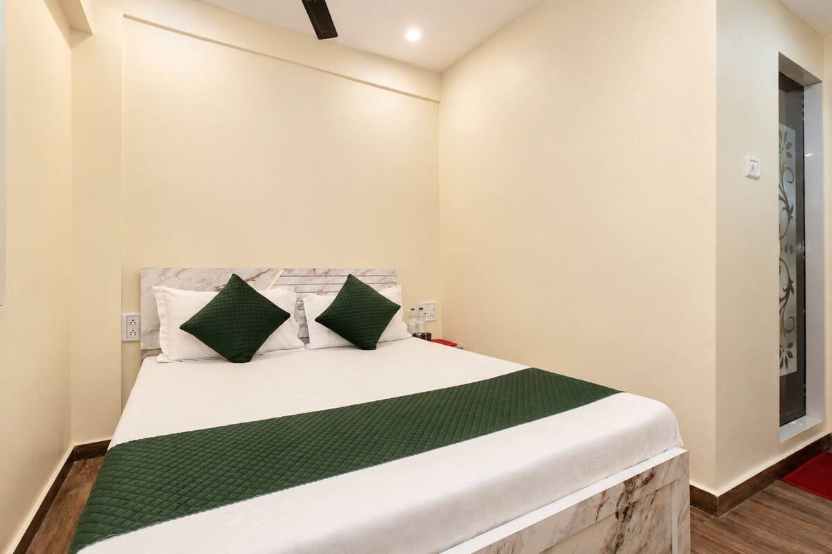 Una cama bien tendida con colcha verde, almohadas blancas y cabecera de mármol en una habitación con paredes color crema y piso de madera.