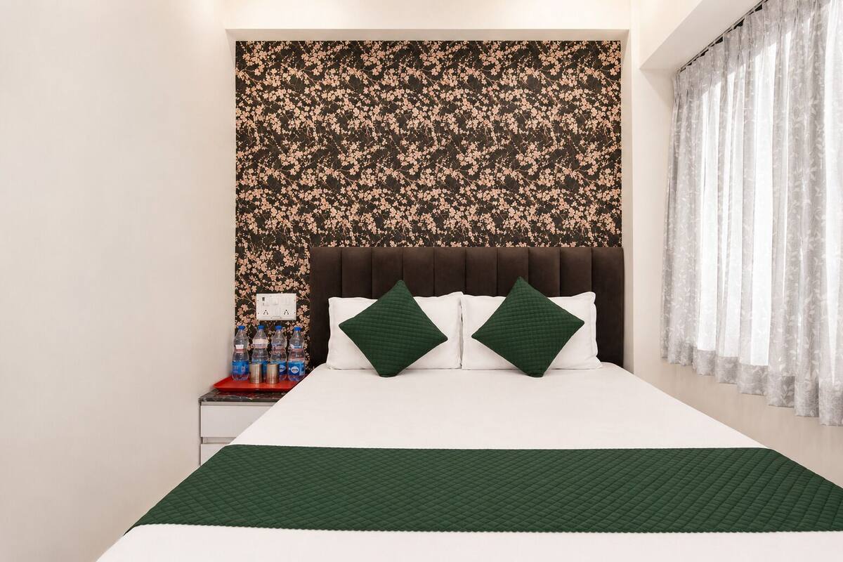 Una habitación de hotel con una cama, almohadillas verdes, papel tapiz floral, una mesita de noche con botellas de agua y una ventana con cortinas de encaje.