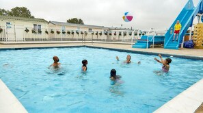 A swimming pool with people playing, a slide, and a building with a white fence.