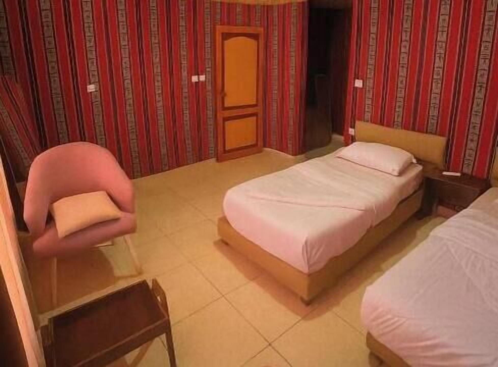Una cama individual con cabecera de madera, una mesita de noche, una silla con cojín y una puerta en una habitación con papel tapiz a rayas.