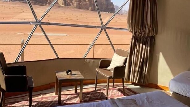 A room with a bed, two chairs, a small table, and a large window offering a view of rocky desert terrain. - Horizon Star Golden Camp (Wadi Rum)