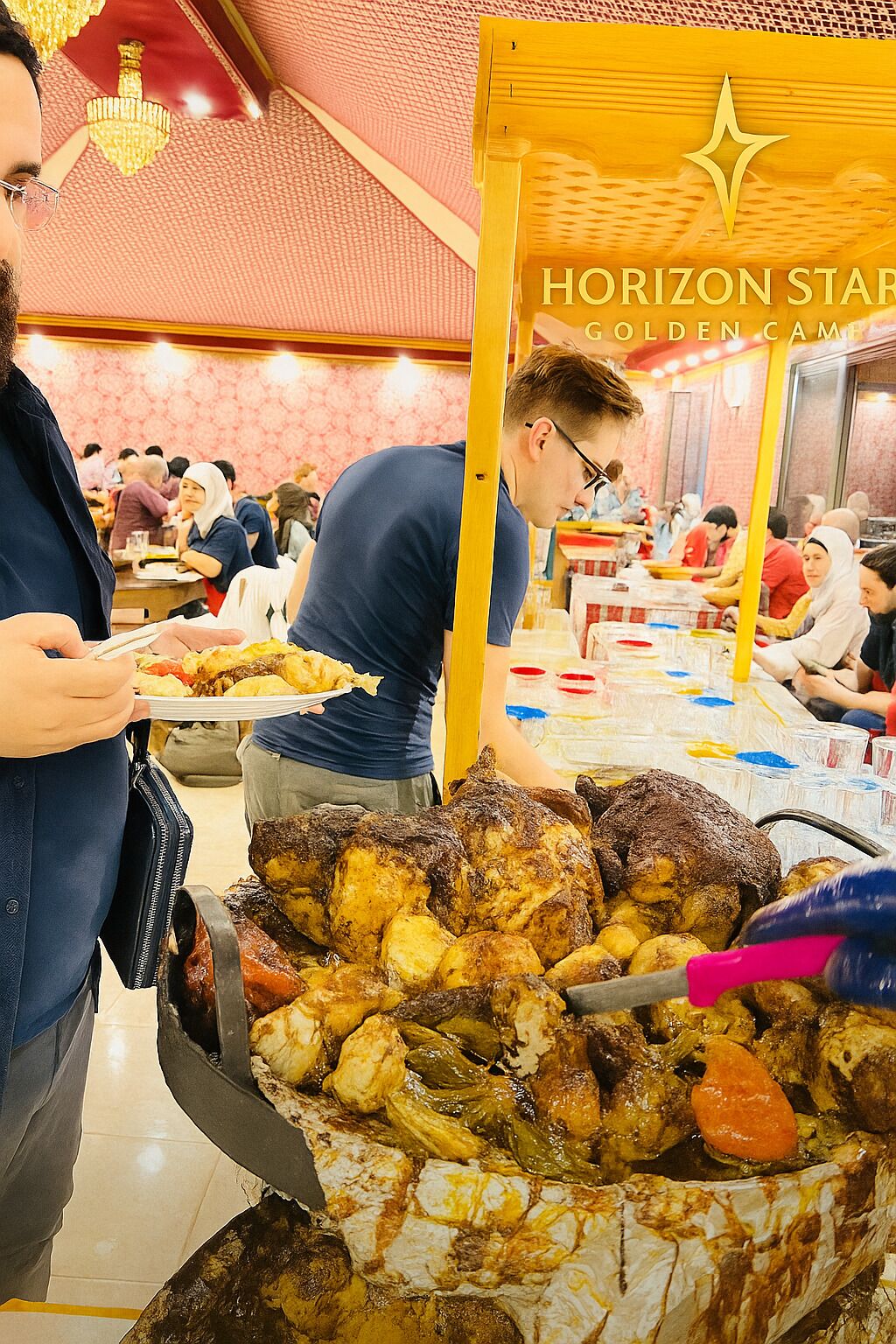 A food stall with roasted meats and vegetables, a Horizon Star sign, and people dining in the background.