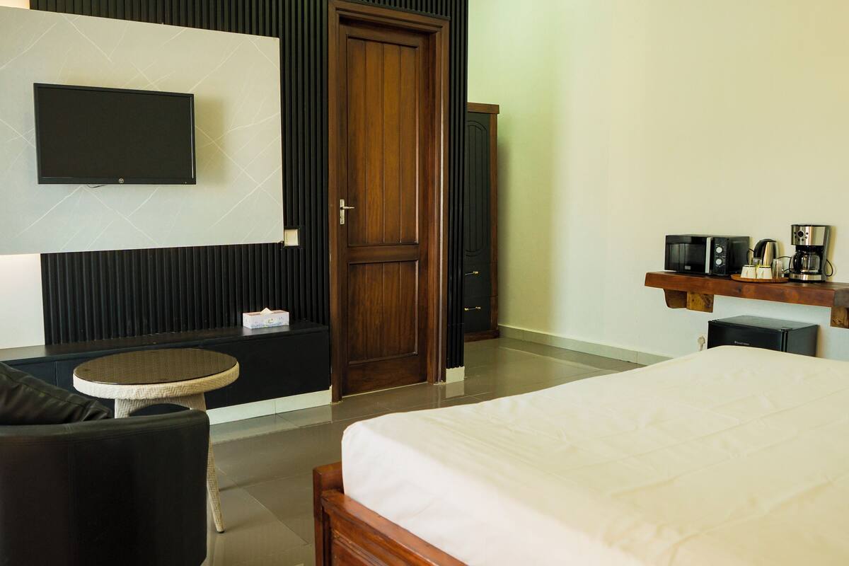 Habitación de hotel con cama, mesa redonda, sofá, televisor montado en la pared, puerta de madera y estante con electrodomésticos.