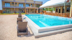 Una piscina con sillas de descanso, una zona de estar cubierta y un edificio con varias ventanas y balcones.